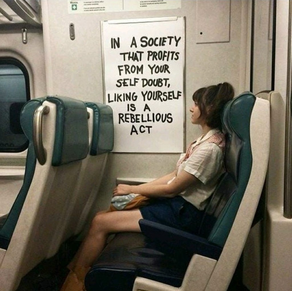 Eine Person sitzt in einem Zug, an der Wand hängt ein Whiteboard mit dem Text 'In a society that profits from your self doubt, liking yourself is a rebellious act