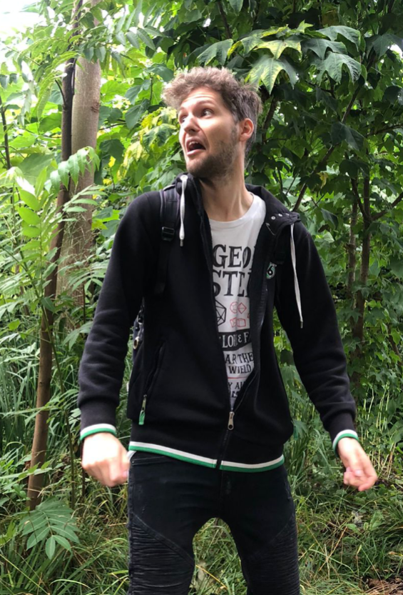 Foto eines jungen Mannes vor grünen Pflanzen; der Mann ist etwas zur Seite gedreht und der Mund ist geöffnet, als würde der Mann erschrocken sein und etwas rufen